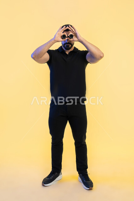 التخطيط المستقبلي للأهداف والطموحات، بورتريه لشاب عربي خليجي اماراتي يرتدي الزي الكاجوال ينظر من خلال المنظار ثنائي العينين، رؤية الاشياء البعيدة عبر التلسكوب، ايماءات وتعابير الدهشة والمفاجئة، حب المغامرة والاستكشاف، صورة الجسم بالطول الكامل، خلفية صفراء