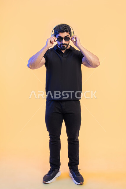 Using headphone techniques, enjoying leisure time, the concept of listening and listening, a portrait of a young Arab Gulf Emirati man wearing casual clothes and sunglasses, wearing headphones, interacting with musical melodies, full-length body portrait, yellow background.