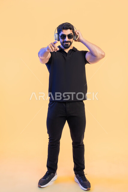 Listening to music enthusiastically, the concept of leisure and entertainment, using headphone technology, a side portrait of a young Arab Gulf Emirati man wearing casual clothes, wearing sunglasses and headphones and pointing to the camera, full-length body portrait, yellow background