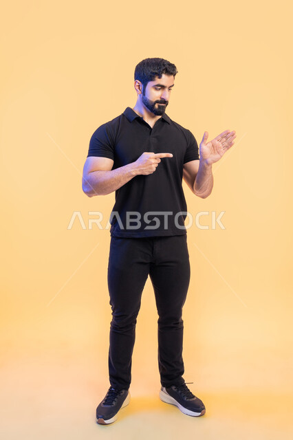 حركات ودلالات مختلفة، تحديد شيء ما، بورتريه لشاب عربي خليجي اماراتي يرتدي الزي الكاجوال ويشير باصبعه السبابة الى كفه، بايماءات السرور، ايماءات التركيز والجدية، صورة الجسم بالطول الكامل، خلفية صفراء