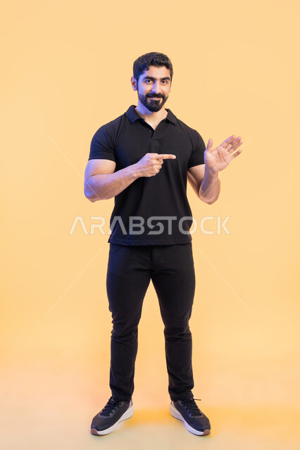 تحديد شيء ما، حركات ودلالات مختلفة، بورتريه لشاب عربي خليجي اماراتي يرتدي زي الكاجوال ويشير باصبعه السبابة على كفه، النظر الى الكاميرا بايماءات السرور، صورة الجسم بالطول الكامل، خلفية صفراء