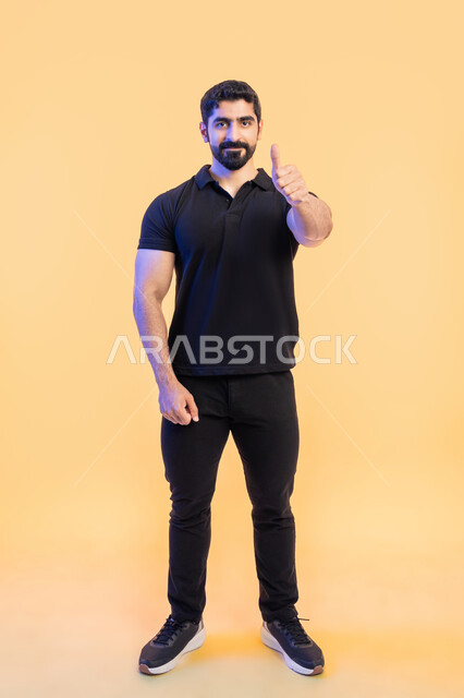 النظر إلى الكاميرا بايماءات القبول والموافقه, الشعور بالإعجاب والرضا والسعاده، بورتريه لشاب عربي خليجي اماراتي مبتسم يرتدي الزي الكاجوال ويرفع أصبع الابهام بعلامة الجودة ،صورة الجسم بالطول الكامل، خلفية رمادية .