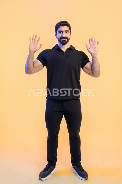 Asking for rescue and assistance, playing, submitting and surrendering, a portrait of a young Arab Gulf Emirati man wearing...  Casual outfit raising hands up with joy and happiness gestures, full body portrait, yellow background