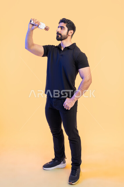 تسجيل مقطع صوتي غنائي، الاستمتاع بأداء المواهب الصوتية، بورتريه من الجانب لشاب عربي خليجي اماراتي يرتدي الزي الكاجوال يمسك بيده المايكروفون، إيماءات وتعابير الاندماج والتفاعل مع الموسيقا، تقديم العروضات المسرحية، صورة الجسم بالطول الكامل، خلفية صفراء