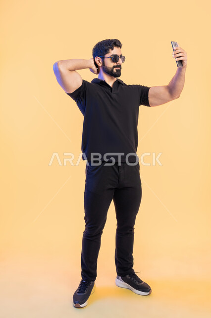 التقاط صور سيلفي تذكارية عبر الجوال، ، استخدام الأجهزة والتقنيات الحديثة، بورتريه لشاب عربي خليجي اماراتي يرتدي الزي الكاجوال والنظارة الشمسية يقوم باجراء مكالمة فيديو عبر تطبيقات ومواقع السوشال ميديا عبر الهاتف المحمول، صورة الجسم بالطول الكامل، خلفية صفراء