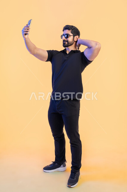 استخدام الأجهزة والتقنيات الحديثة، التقاط صور سيلفي تذكارية عبر الجوال، بورتريه لشاب عربي خليجي اماراتي يرتدي الزي الكاجوال والنظارة الشمسية يقوم باجراء مكالمة فيديو عبر تطبيقات ومواقع السوشال ميديا عبر الهاتف المحمول، صورة الجسم بالطول الكامل، خلفية صفراء