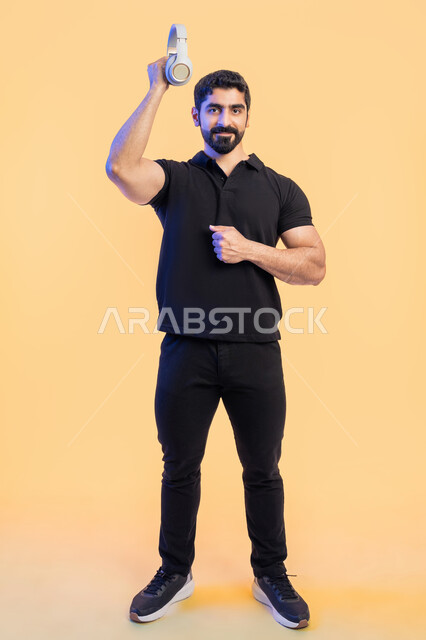 استخدام تقنية الهيدفون، بورتريه لشاب عربي خليجي اماراتي مبتسم يرتدي الزي الكاجوال يرفع سماعة الرأس إلى الأعلى بتعابير السرور، قضاء أوقات مسلية في أوقات الفراغ، الاستمتاع للمواهب الغنائية، صورة الجسم بالطول الكامل، خلفية صفراء