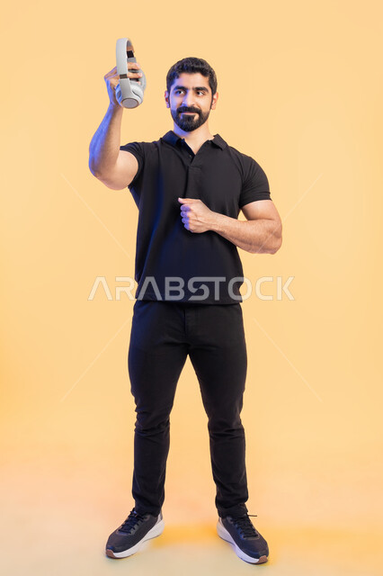 قضاء أوقات مسلية في أوقات الفراغ، استخدام تقنية الهيدفون، بورتريه لشاب عربي خليجي اماراتي مبتسم يرتدي الزي الكاجوال يرفع سماعة الرأس إلى الأعلى بتعابير السرور، الاستمتاع للمواهب الغنائية، صورة الجسم بالطول الكامل، خلفية صفراء