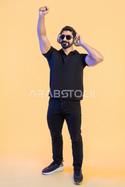 التفاعل والاندماج مع الانغام الموسيقية، بورتريه لرجل عربي خليجي اماراتي يرتدي الزي الكاجوال والنظارات الشمسية يضع سماعة الرأس، الاستماع للأغاني باسخدام الهيدفون بتعابير الرقص، صورة الجسم بالطول الكامل, خلفية صفراء
