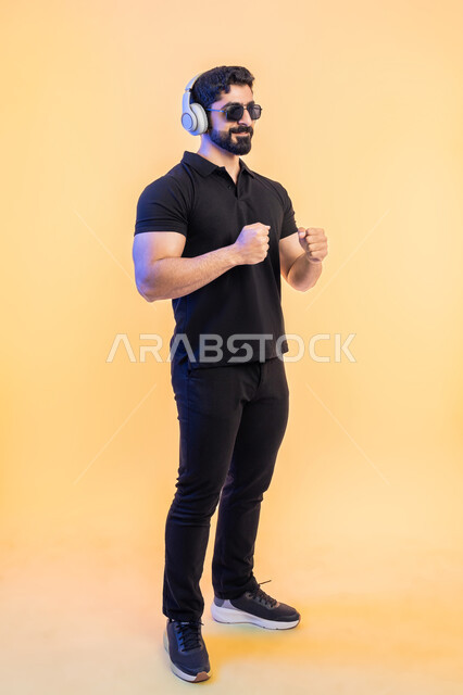 الاستماع للأغاني باسخدام الهيدفون، بورتريه من الجانب لرجل عربي خليجي اماراتي يرتدي الزي الكاجوال والنظارات الشمسية يضع سماعة الرأس، التفاعل والاندماج مع الانغام الموسيقية، صورة الجسم بالطول الكامل, خلفية صفراء