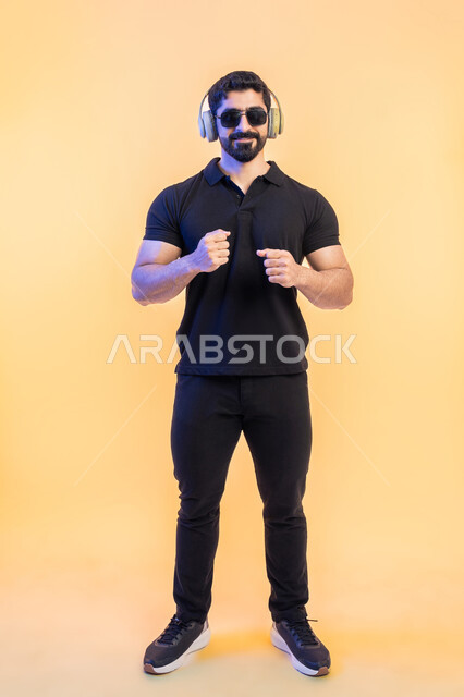 إيماءات وتعابير الاندماج مع الموسيقى، بورتريه لرجل عربي خليجي اماراتي يرتدي الزي الكاجوال والنظارات الشمسية يضع سماعة الرأس، الاستماع للأغاني باسخدام الهيدفون، التفاعل مع الانغام الموسيقية، صورة الجسم بالطول الكامل, خلفية صفراء