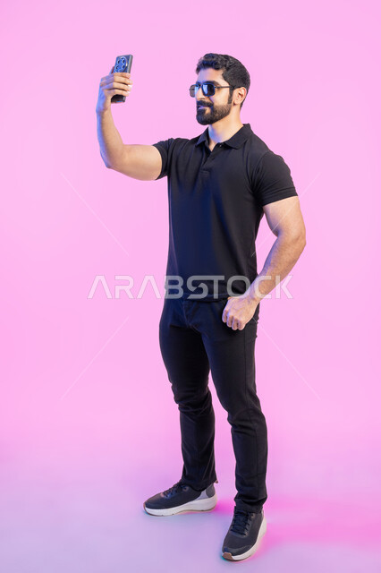التقاط صور سيلفي تذكارية عبر الجوال، ، استخدام الأجهزة والتقنيات الحديثة، بورتريه لشاب عربي خليجي اماراتي يرتدي الزي الكاجوال والنظارة الشمسية يقوم باجراء مكالمة فيديو عبر تطبيقات ومواقع السوشال ميديا عبر الهاتف المحمول، صورة الجسم بالطول الكامل، خلفية وردية