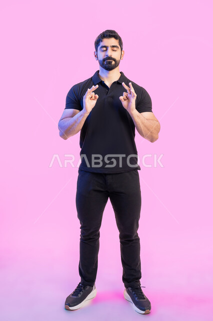 الشعور بالرضا و السعادة, حركات الاعجاب والقبول, بورتريه لشاب عربي خليجي اماراتي يرتدي الزي الكاجوال يرفع يده للأعلى بإيماءات علامة الجودة و السرور, صورة الجسم بالطول الكامل، خلفية وردية
