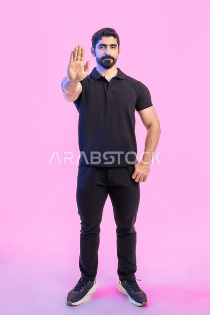 Standing in anger and rejecting a specific order, asking not to interfere and lower the voice and threats, silence and stop talking, refraining from doing a specific order, a portrait of a young Arab Gulf Emirati man wearing a casual outfit, raising his palm up with gestures of annoyance, full-length body portrait, pink background.