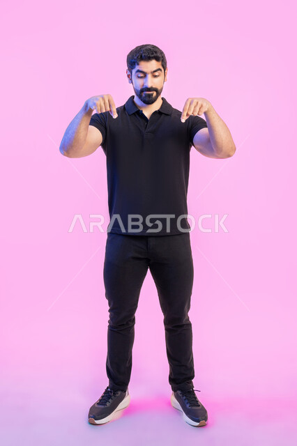 حركات تدل على هذا المكان (هنا)، تحديد موقع معين، بورتريه لشاب عربي خليجي اماراتي يرتدي الزي الكاجوال الأسود  يشير بأصابع السبابة نحوالأسفل، في هذه اللحظة (الآن)، النظر إلى شيء ما، صورة الجسم بالطول الكامل، خلفية وردية
