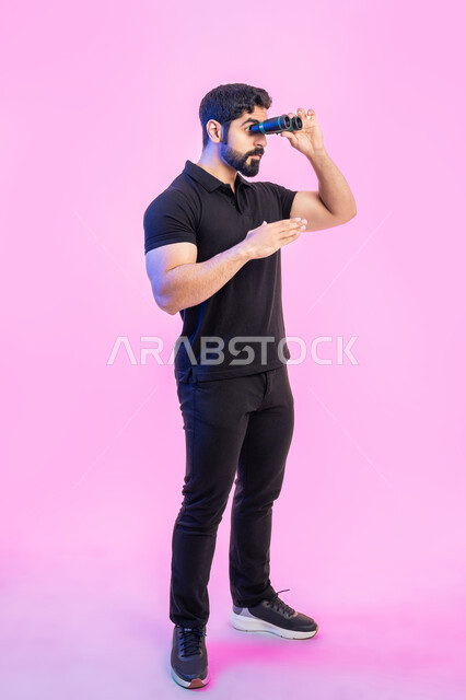 التطلع والنظرة المستقبلية, مفهوم المغامرة والاستكشاف, بورتريه لشاب عربي خليجي اماراتي يرتدي الزي الكاجوال ينظر من خلال منظار الرؤية ويشير بيده إلى الأمام, رؤية الأشياء البعيدة عبر ناظور ثنائي العينين التلسكوب, صورة الجسم بالطول الكامل، خلفية وردية