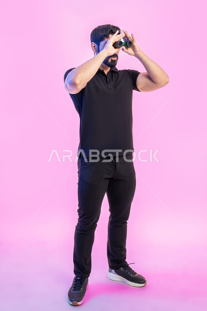 طموحات وأهداف مستقبلية، البحث عن شيء ما وتوسيع مجال الرؤية باستخدام المنظار، مفهوم الاستكشاف والمغامرة، بورتريه لشام عربي خليجي اماراتي يرتدي الزي الكاجوال يمسك بيده التلسكوب ثنائي العينين، صورة الجسم بالطول الكامل، خلفية وردية