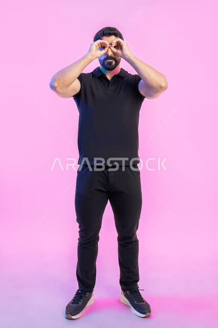 التخطيط المستقبلي للأهداف والطموحات، بورتريه لشاب عربي خليجي اماراتي يرتدي الزي الكاجوال يضع يديه على عينه على شكل المنظار ثنائي العينين، حب المغامرة والاستكشاف، تعابيرالاندماج والجدية، رؤية الاشياء البعيدة، صورة الجسم بالطول الكامل، خلفية وردية