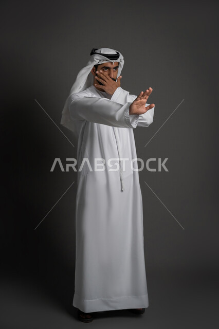 الصمت والتوقف عن الكلام, الوقوف بغضب ورفض أمر محدد, بوتريه لشاب عربي خليجي إماراتي يرتدي الكندورة والغنرة يرفع كفه للأعلى بإيماءات الانزعاج ويغطي جزء من وجهه, طلب عدم التدخل والامتناع عن فعل أمر معين, صورة لكامل الجسم، خلفية سوداء