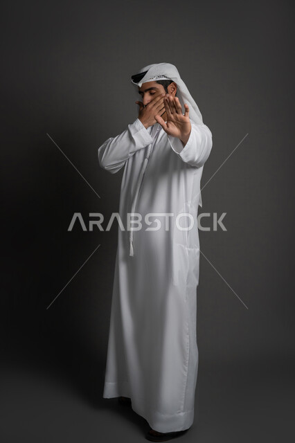 Standing in anger and refusing a specific order, asking not to interfere and refrain from doing a specific order, being silent and stopping talking, a portrait of a young Arab Gulf Emirati man wearing a kandura and a ghunra raising his palm up in gestures of annoyance and covering part of his face, a full body image, black background