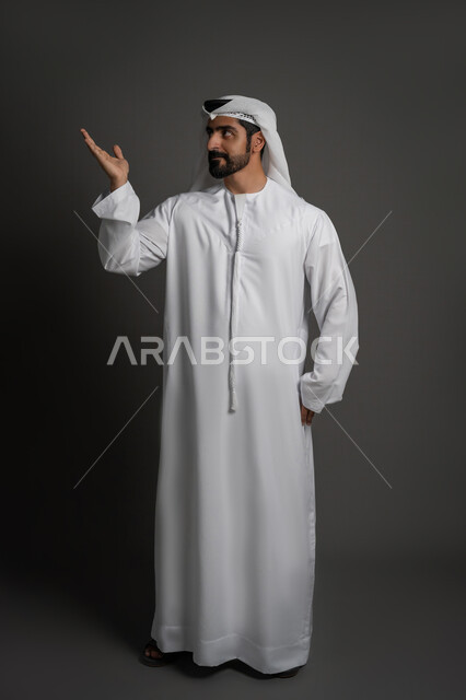 الإشارة إلى مكان ما، تحديد موقع معين, بورتريه لرجل عربي خليجي اماراتي يرتدي الكندورة والغترة البيضاء ينظر ويشيربيده نحو اتجاه واحد, صورة لكامل الجسم، خلفية سوداء