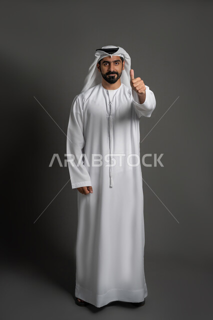 مفهوم الأناقة والرجولة والاهتمام بالمظهر الخارجي, بورتريه لرجل عربي خليجي إماراتي يرتدي الكندورة البيضاء والغترة ويرفع اصبع الابهام بإيماءة إلى الموافقة والقبول, الوقوف باستقامة والنظر إلى الكاميرا بتعابير الثقة بالنفس, صورة لكامل الجسم، خلفية سوداء