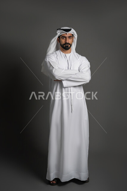 The concept of masculinity, strength and self-confidence, a portrait of an Arab Gulf Emirati man wearing a kandura and a ghutra, looking at the camera with gestures of seriousness and firmness, standing with folded hands, interest in elegance and external appearance, a full body image, black background