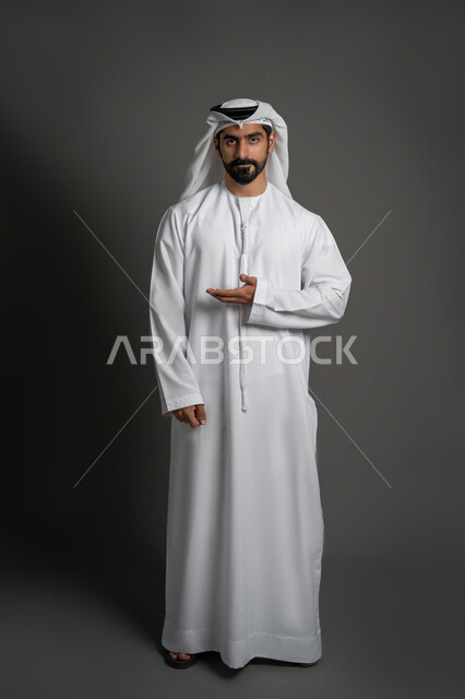 النظر إلى الكاميرا بإيماءات الحزم والجدية، الاشارة الى شيء ما، بورتريه لرجل عربي خليجي إماراتي يرتدي الكندورة والغترة البيضاء يقوم بفتح كفه ويشير إلى اتجاه معين، صوره الجسم بالطول الكامل، خلفيه سوداء