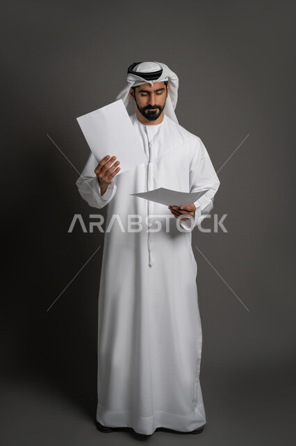 النظر الى الملفات بإيماءات التركيز والتدقيق، بورتريه لرجل عربي خليجي اماراتي يرتدي الكندورة والغترة يحمل بيديه اوراق بيضاء، متابعة المراسلات والسجلات، انجاز المهام المطلوبة،  صورة لكامل الجسم، خلفيه سوداء