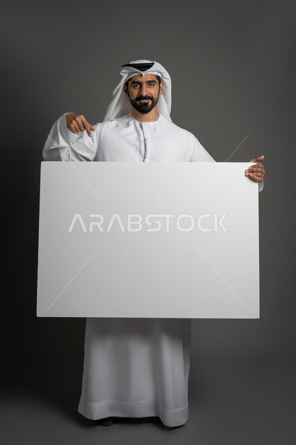 مفهوم الإعلان والتسويق، استخدام لوحات توضيحية والإشارة اليها، لوح إعلاني فارغ، موك اب أبيض، بورتريه لرجل عربي خليجي إماراتي مبتسم يرتدي الكندورة والغترة يحمل بيده لوحه بيضاء مستطيلة كبيرة وينظر إلى الكاميرا بإيماءات الثقة بالنفس، صورة لكامل الجسم، خلفية سوداء