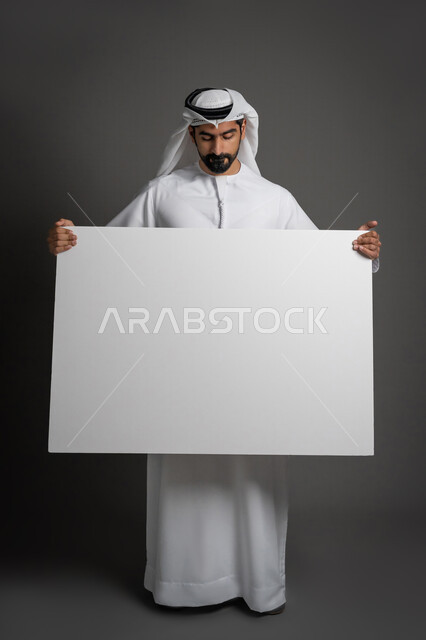 موك اب أبيض، لوح إعلاني فارغ، مفهوم الإعلان والتسويق، استخدام لوحات توضيحية، بورتريه لرجل عربي خليجي إماراتي مبتسم يرتدي الكندورة والغترة يحمل بيده لوحه بيضاء مستطيلة كبيرة وينظر إليها، صورة لكامل الجسم، خلفية سوداء