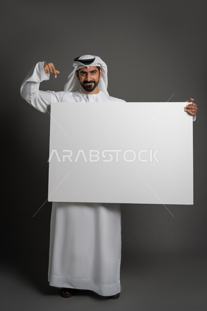 استخدام لوحات توضيحية والإشارة اليها، مفهوم الإعلان والتسويق، لوح إعلاني فارغ، موك اب أبيض، بورتريه لرجل عربي خليجي إماراتي مبتسم يرتدي الكندورة والغترة يحمل بيده لوحه بيضاء مستطيلة كبيرة وينظر إلى الكاميرا بإيماءات البهجة والسرور، صورة لكامل الجسم، خلفية سوداء
