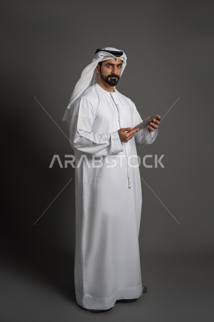 النظر الى الكاميرا بتعابير الحزم والجدية، إجراء مكالمات الفيديو، استخدام الأجهزة الحديثة في التواصل مع الأصدقاء, بورتريه لرجل عربي خليجي اماراتي يرتدي الكندورة والغترة البيضاء يمسك بيده جهاز التابليت، صورة لكامل الجسم، خلفية سوداء