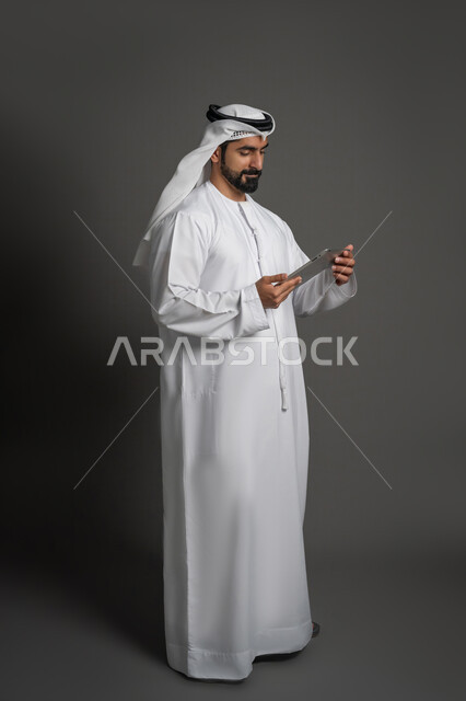 إيماءات وتعابير الإندماج أثناء إجراء مكالمة فيديو، استخدام الأجهزة الحديثة في التواصل مع الأصدقاء, بورتريه لرجل عربي خليجي اماراتي يرتدي الكندورة والغترة البيضاء يمسك بيده جهاز التابليت، صورة لكامل الجسم، خلفية سوداء