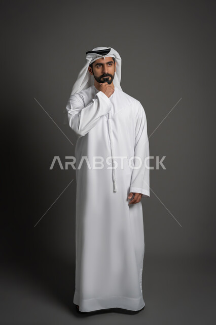 Planning for the future and setting goals, looking at something with focused expressions, portrait of an Arab Gulf Emirati man wearing a white kandura and ghutra putting his hand on his chin with thinking gestures, full body photo, black background
