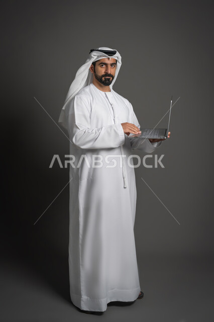 Looking at the camera with serious gestures, using modern devices to accomplish tasks, portrait of an Arab Gulf Emirati man wearing a white kandura and ghutra clicking on the computer converter, full body image, black background