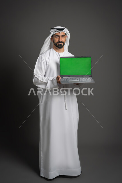 عرض شاشة خضراء عبر اللابتوب, استخدام الأجهزة الحديثة في انجاز المهام, بورتريه لرجل عربي خليجي اماراتي يرتدي الكندورة والغترة البيضاء يحمل الحاسوب المحمول يقوم بالإشارة إليه، النظر إلى الكاميرا بإيماءات الجدية، صورة لكامل الجسم، كروما، خلفية سوداء
