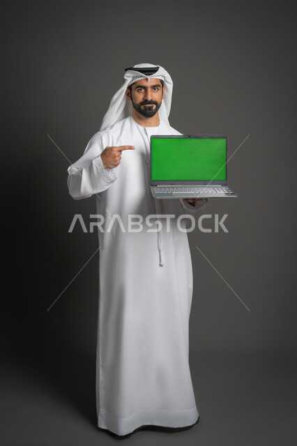 استخدام الأجهزة الحديثة في انجاز المهام, بورتريه لرجل عربي خليجي اماراتي يرتدي الكندورة والغترة البيضاء يحمل الحاسوب المحمول يقوم بالإشارة إليه، عرض شاشة خضراء عبر اللابتوب, النظر إلى الكاميرا بإيماءات الجدية، صورة لكامل الجسم، كروما، خلفية سوداء