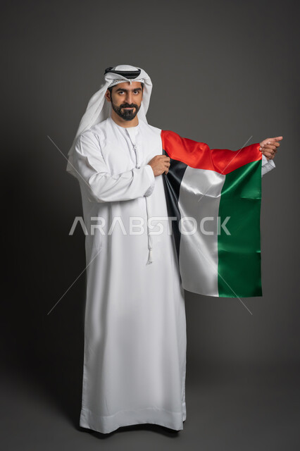 الإحتفال بالعيد الوطني الإماراتي، النظر إلى الكاميرا بتعابير الفخر، إحياء ذكرى الإستقلال لدولة الإمارات العربية المتحدة، بورتريه لرجل عربي خليجي إماراتي يرتدي الغترة والكندورة الإماراتية يحمل علم الإمارات، صورة لكامل الجسم، خلفية سوداء
