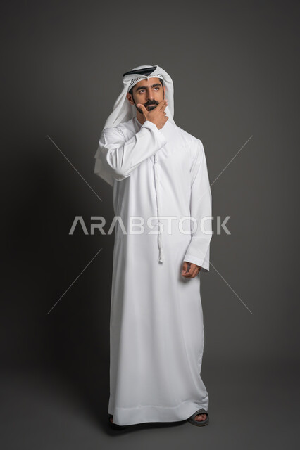 التخطيط للمستقبل ووضع الأهداف، النظر إلى شيء ما بتعابير التركيز، بورتريه لرجل عربي خليجي إماراتي يرتدي الكندورة والغترة البيضاء يضع يده على ذقنه بإيماءات التفكير, صورة لكامل الجسم، خلفية سوداء