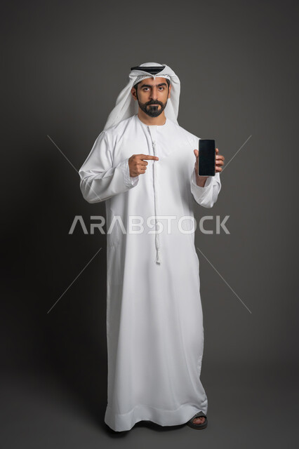 التسويق الإلكتروني وعرض خدمات وتطبيقات الجوال، بورتريه لرجل عربي خليجي إماراتي يرتدي الكندورة والغترة البيضاء ويشير بإصبع السبابة نحو الهاتف المحمول بشاشة سوداء فارغة، إستخدام برامج الهاتف الذكي، صورة لكامل الجسم، خلفية سوداء