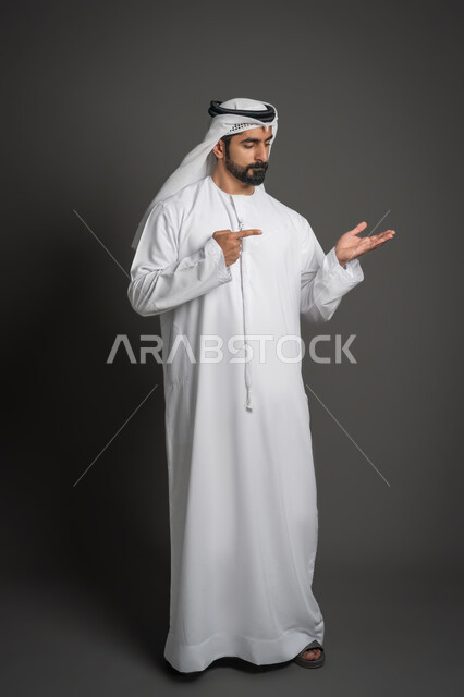 إيماءات حمل ورفع شيء ما، بورتريه لرجل عربي خليجي إماراتي يرتدي الكندورة الإماراتية والغترة البيضاء يشير باصبع السبابة ليده، الإشارة نحو هدف ما، حركة أصابع وإشارات، صورة لكامل الجسم، خلفية سوداء