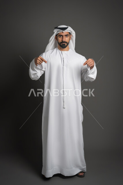 تحديد موقع معين, اشارات تحدد هذا المكان (هنا)، حركات وتعابير وجود شيء ما، بورتريه لرجل عربي خليجي اماراتي يرتدي الغترة والكندورة يشير بأصابعه نحو الأسفل, النظر إلى الكاميرا بإيماءات السرور، صورة لكامل الجسم، خلفية سوداء