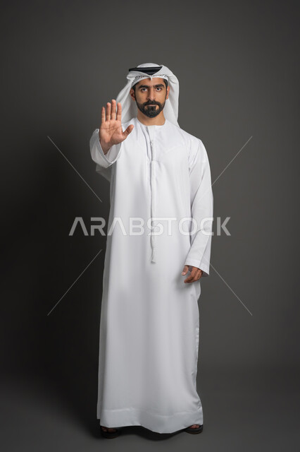 النظر إلى الكاميرا بتعابير الحزم والجدية، طلب المنع والتوقف عن فعل شيء ما، بورتريه لرجل عربي خليجي إماراتي يرتدي الكندورة والغترة يرفع يده بإيماءات الرفض والامتناع، صورة لكامل الجسم، خلفية سوداء