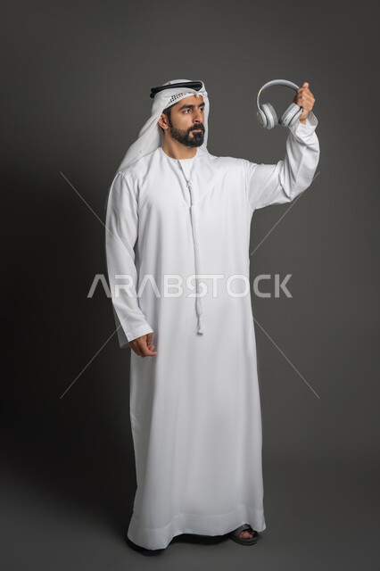 تقنيات حديثة وتكنولوجيا, سماعة الاتصال وسماع الموسيقا, استخدام تقنية الهيدفون, بورتريه لرجل عربي خليجي اماراتي يرتدي الكندورة والغترة يمسك بيده سماعات الأذن اللاسلكية ينظر إليها بإيماءات الحزم والجدية, صورة لكامل الجسم، خلفية سوداء