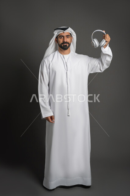 Using headphone technology, headset for communication and listening to music, portrait of an Arab Gulf Emirati man wearing a kandura and ghutra holding wireless earphones, modern techniques and technology, full body image, black background