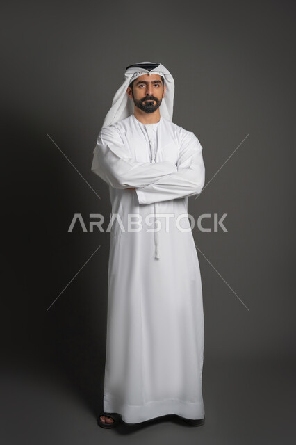 مفهوم الرجولة والقوة والثقة بالنفس، بورتريه لرجل عربي خليجي إماراتي يرتدي الكندورة والغترة ينظر الى الكاميرا بإيماءات الجدية والحزم يقف بأيدي مكتوفة، الاهتمام بالأناقة والمظهر الخارجي، صورة لكامل الجسم، خلفية سوداء