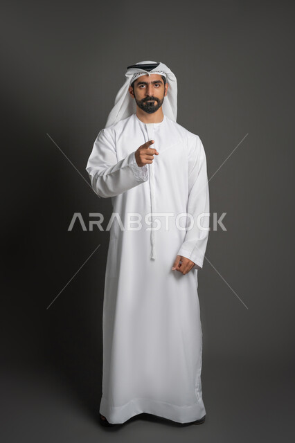 الاشارة إلى شيء ما, الوقوف باستقامة والنظر الكاميرا بإيماءات الحزم والجدية, بورتريه لرجل عربي خليجي إماراتي يرتدي الكندورة والغترة البيضاء يشير بإصبع السبابة نحو مكان معين, صورة الجسم بالطول الكامل, خلفية سوداء