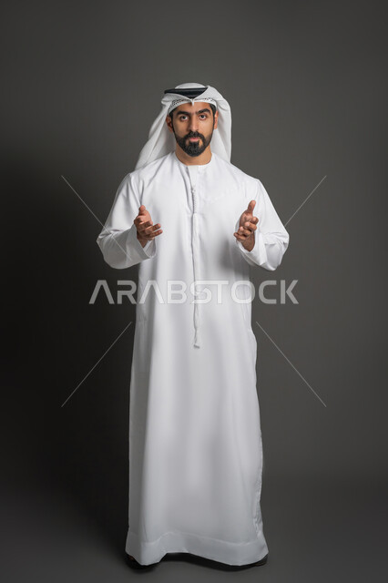 الاهتمام بالمظهر الخارجي، بورتريه لرجل عربي خليجي إماراتي يرتدي الكندورة والغترة البيضاء يوجه كفيه باتجاه الكاميرا بإيماءات الترحيب، مفهوم الاناقه والرجولة، الإشارة إلى شيء ما، صورة لكامل الجسم، خلفية سوداء