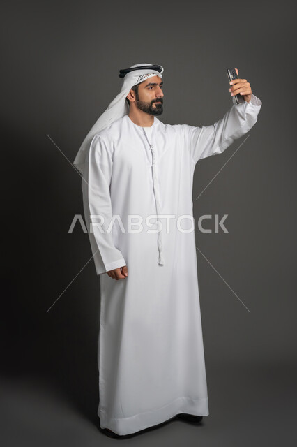 إجراء مكالمة فيديو بإستخدام الهاتف المحمول، بورتريه لرجل عربي خليجي إماراتي يرتدي الكندورة والغترة يقوم باستعمال الأجهزة التقنية الحديثة في التواصل مع الأصدقاء، صورة لكامل الجسم، خلفية سوداء